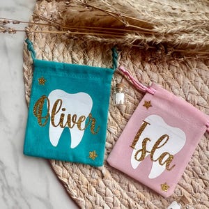 May include: Two small fabric drawstring pouches, one teal and one light pink, each featuring a white tooth graphic and a name in gold glitter script. The teal pouch says Oliver and the pink pouch says Isla, both accented with small gold glitter stars.