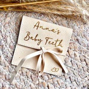 May include: A beige suede pouch tied with a matching ribbon. The pouch is embroidered with the words "Anna's Baby Teeth" in gold script. Two small gold hearts are embroidered below the text. The pouch is on a woven surface.