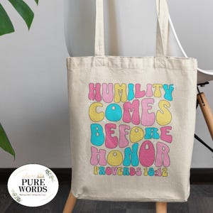 May include: A beige canvas tote bag featuring the text "HUMILITY COMES BEFORE HONOR PROVERBS 18:12" in colourful bubble letters. The bag has two handles and a round logo in the lower left corner.