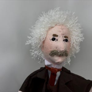 Puede incluir: Una muñeca de tela con tez pálida, una peluca de lana blanca y un bigote de lana marrón. La muñeca viste una camisa blanca con cuello, una chaqueta marrón oscuro y una corbata roja fina. Los rasgos faciales incluyen ojos de cuentas negras.