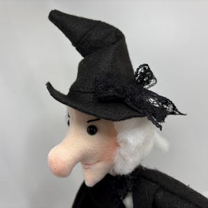 Puede incluir: Una muñeca de fieltro con rostro pálido, nariz prominente, ojos de cuentas negras y cabello blanco. La muñeca lleva un sombrero negro puntiagudo adornado con un lazo de encaje negro. Viste una prenda negra con un detalle de cuello de encaje.