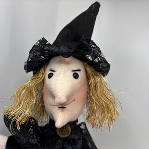 Puede incluir: Una muñeca de tela que representa a una bruja con un sombrero negro puntiagudo adornado con encaje negro. La muñeca tiene piel clara, una nariz prominente, rasgos faciales pintados y cabello rubio sintético. Se observa un pequeño botón dorado.