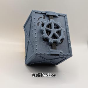 Könnte beinhalten: Eine graue, kastenförmige Deckbox mit einem zahnradförmigen Schloss. Die Box hat ein genietetes Design und den Text "Vault Deckbox" unten. Das Zahnrad ist mittig auf der Vorderseite angebracht.