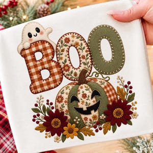 Pode incluir: Um artigo de tecido branco com um design bordado exibindo a palavra BOO. O B é xadrez castanho, o O é floral e o segundo O é verde sólido. Um pequeno fantasma branco está sobre o B e uma abóbora com detalhes florais está abaixo.