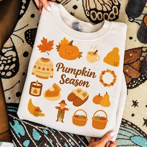 May include: A light beige crewneck sweatshirt featuring the text Pumpkin Season in brown script. The graphic includes illustrations of a smiling pumpkin, a sweater, a mug, a beanie, a candle, pie, slippers, a cornucopia, a scarecrow, and baskets.