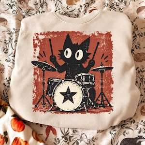 May include: A beige crewneck sweatshirt featuring a graphic print of a black cat playing a drum set. The cat has large white eyes and holds drumsticks. The bass drum is decorated with a black star. The background of the print is a distressed red square.