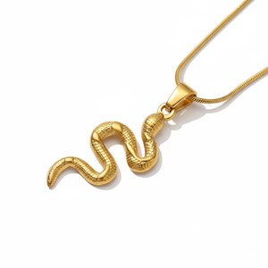 May include: A gold-coloured snake pendant necklace. The pendant is a detailed, three-dimensional snake with textured scales. The necklace chain is a thin, gold-coloured cable chain. The jewellery is displayed against a white background.
