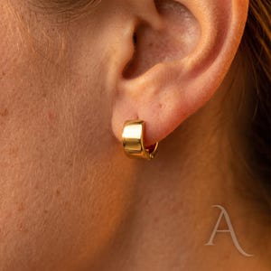 Peut inclure: Gros plan d'une boucle d'oreille créole dorée portée à l'oreille. La boucle d'oreille a un design épais et arrondi et une finition brillante. La peau autour de l'oreille est visible, avec un ton chaud. La boucle d'oreille est le point central, mettant en valeur son style simple mais élégant.