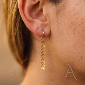 May include: A gold hoop earring featuring a long, dangling charm composed of a vertical chain of small, detailed leaf-shaped metal links. The earring is worn in the earlobe, showcasing a polished metallic finish and a delicate, symmetrical botanical design.