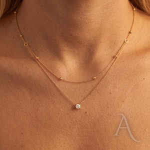 May include: A double-layered gold necklace featuring a delicate chain with small evenly spaced gold beads on the upper strand and a single round clear gemstone pendant centred on the lower strand, resting against the collarbone area.