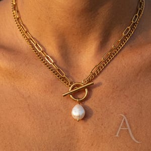 May include: Gold-coloured necklace with two layered chains. The top chain is a fine curb chain, the lower one a larger link chain. A circular clasp with a bar closure holds a white, pear-shaped pearl pendant.