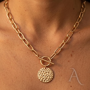 May include: Gold-coloured necklace with a chunky chain and a hammered disc pendant. The necklace has a toggle clasp. The chain links are oval. The pendant is round with a textured surface.