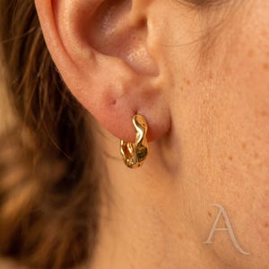 May include: A small, gold-toned hoop earring with a wavy, irregular surface texture. The earring is circular in shape and features a polished, reflective finish that catches the light, creating subtle highlights along its curved, undulating edges.