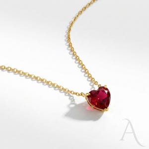 May include: A gold necklace featuring a heart-shaped, red gemstone pendant. The pendant is faceted and set in a gold bezel. The chain is fine, and the necklace is shown against a white backdrop.