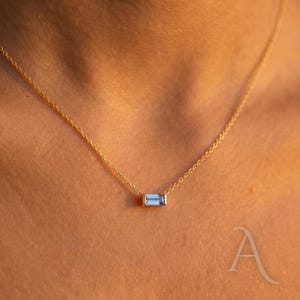 Peut inclure: Un collier en or délicat avec un pendentif rectangulaire en pierre précieuse bleu clair. Le pendentif est suspendu à une fine chaîne en or, créant un design minimaliste et élégant. Le collier est présenté sur un fond de teint.