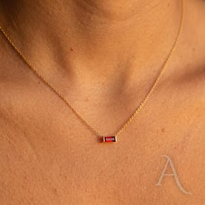 May include: A gold necklace with a rectangular, red gemstone pendant. The pendant is set in a gold frame and hangs from a fine gold chain. The necklace is shown against a skin tone background.