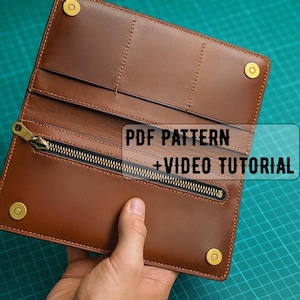 Op de afbeelding: Open bruin leren portemonnee met een ritsvak en drukknoopsluitingen. De portemonnee heeft zichtbare stiksels en goudkleurige hardware. Tekst op de afbeelding luidt "PDF PATTERN + VIDEO TUTORIAL".