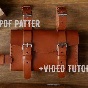 以下が含まれることがあります： 調節可能なストラップとシルバーのバックルが付いた茶色の革製バッグ。バッグはフラップ開閉式で、より大きなオブジェクトに取り付けるように設計されています。画像には「PDF PATTER」と「+ VIDEO TUTORIAL」と書かれています。