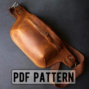 Op de afbeelding: Een bruine leren heuptas met ritssluiting en verstelbare riem. De tas heeft een rechthoekige vorm met afgeronde hoeken en een zilverkleurige metalen ring. De tekst "PDF PATTERN" wordt weergegeven in een witte rechthoek.