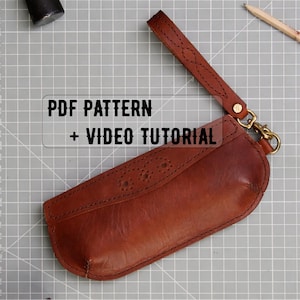 May include: A handcrafted brown leather glasses case with a wrist strap, featuring decorative stitching and a brass clasp. The case is shown on a gray cutting mat, with the text "PDF PATTERN + VIDEO TUTORIAL".