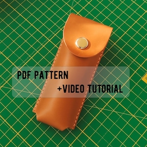 以下が含まれることがあります： 金色のスナップで留められたフラップクロージャー付きのタンレザーシース。シースは長方形で上部が丸く、緑色のカッティングマットの上に置かれています。画像には「PDF PATTERN + VIDEO TUTORIAL」と書かれています。