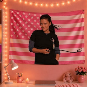 Könnte beinhalten: Eine rosa-weiße amerikanische Flagge hängt an einer rosa Wand. Eine Person in einem schwarzen Oberteil hält ein Stethoskop vor der Flagge. Der Raum ist mit Lichterketten, einer Schreibtischlampe und einem Schreibtisch mit einem Laptop dekoriert.