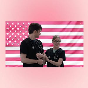 May include: A pink American flag background with two people in black scrubs and stethoscopes. The flag features white stars and pink stripes. The people are smiling and looking at each other.