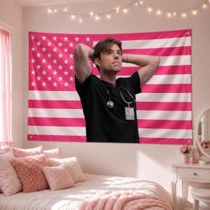 Op de afbeelding: Een roze en witte Amerikaanse vlag wandtapijt met een persoon in een zwarte scrub top en een stethoscoop. Het wandtapijt hangt boven een bed met roze kussens en een roze deken. De kamer is ingericht in roze tinten.