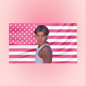 May include: A pink American flag with white stars and pink stripes. A person wearing a white tank top is positioned in front of the flag. The background is a solid pink color.