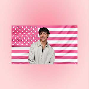 May include: A person wearing a light grey jacket and a black shirt, smiling in front of a pink American flag. The flag has white stars and pink stripes. The background is a solid pink colour.