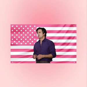 May include: A pink American flag with white stars and stripes. A person wearing a purple button-down shirt stands in front of the flag. The background is a solid pink color.