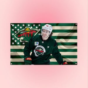 Puede incluir: Una bandera de pared rectangular que muestra a un jugador de hockey con una camiseta verde oscuro con el logotipo de Minnesota Wild y el texto Property of Minnesota. El fondo es una bandera estadounidense estilizada con rayas verdes y crema.