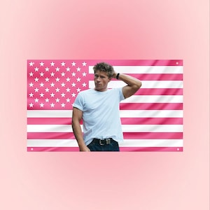 Könnte beinhalten: Eine Person in einem weißen T-Shirt und einer blauen Jeans steht vor einer rosa amerikanischen Flagge. Die Flagge hat weiße Sterne und rosa und weiße Streifen. Der Hintergrund ist einfarbig rosa.