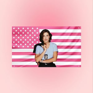 Könnte beinhalten: Eine Frau steht vor einer rosa amerikanischen Flagge. Sie trägt ein hellblaues Hemd, eine schwarze Hose und ein Abzeichen. Eine schwarze Jacke liegt über ihrer Schulter. Die Flagge hat weiße Sterne und rosa Streifen.