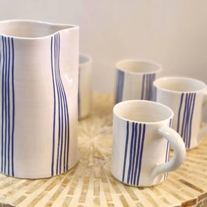 Peut inclure: Un ensemble de pichet et de tasses en céramique blanche avec des rayures bleues verticales. Le pichet a une grande anse et un bec verseur. Les tasses ont des anses et sont disposées autour du pichet.