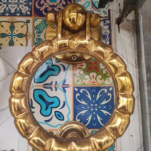 Puede incluir: Un aldaba dorada con un asa en forma de anillo y una placa trasera decorativa. Está montada en una pared con azulejos estampados de colores azul, blanco y verde.