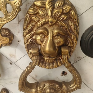 Puede incluir: Un aldaba de puerta de latón con forma de cabeza de león con una melena detallada y un asa de anillo. La aldaba tiene forma ovalada y tiene un diseño floral decorativo en el asa. Se ve otra ferretería de latón.