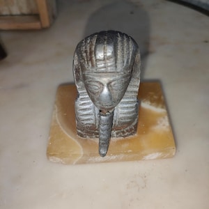 Puede incluir: Una pequeña escultura de metal antigua de una esfinge con una cara y un tocado detallados. La escultura está montada sobre una base rectangular de piedra de color claro. La esfinge tiene una apariencia envejecida.