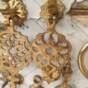 Puede incluir: Primer plano de varios aldabas de latón antiguas. Las aldabas presentan intrincados diseños de estrellas y flores. El latón tiene un tono dorado cálido, y los diseños están detallados con varios patrones y formas. El fondo es una superficie blanca.
