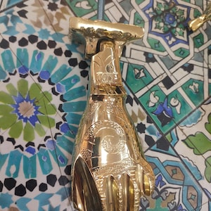 Puede incluir: Un aldaba dorada con forma de mano, con intrincados detalles grabados. La mano sostiene un diseño en espiral. Está unida a una varilla roscada. El fondo es un mosaico de azulejos coloridos.