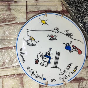 Könnte beinhalten: Weiße Keramikplatte mit blauem Rand, die Cartoon-Skifahrer, Snowboarder und eine Sonne zeigt. Auf der Platte steht in schwarzer Schrift "estuve esquiando en SIERRA NEVADA". Zwei goldfarbene Metallhalter sind angebracht.