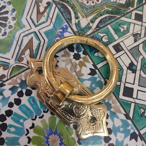 Könnte beinhalten: Ein Messing-Schubladengriff mit Ringgriff und verzierter Rückplatte. Die Beschläge sind goldfarben und liegen auf einer bunten Fliesenoberfläche mit Blau-, Grün- und Weißtönen. Der Schubladengriff ist wahrscheinlich für einen Schrank oder ein Möbelstück.