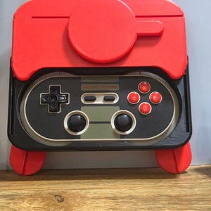 May include: A red and black retro-style game controller with a matching red top. The controller features a directional pad, buttons, and a 'SELECT START' label. The design is reminiscent of classic gaming consoles.