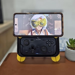Könnte beinhalten: Ein Smartphone mit einer animierten Figur mit goldfarbener Brille liegt auf einem schwarzen Acegamer-Controller. Der Controller hat gelbe Füße und ein Steuerkreuz mit den Tasten A, B, X und Y.