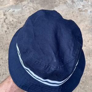 May include: Navy blue bucket hat with two white stripes around the brim. The hat is made of a soft fabric and has a classic, casual style. A simple, versatile accessory for sun protection.