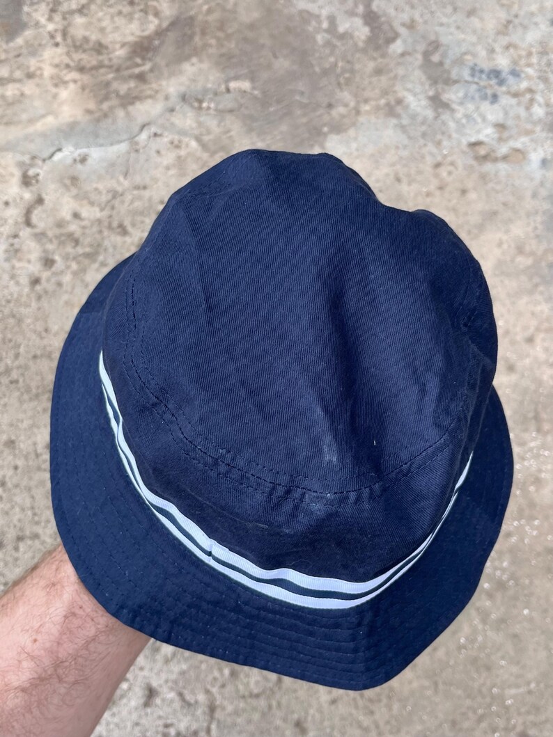 May include: Navy blue bucket hat with two white stripes around the brim. The hat is made of a soft fabric and has a classic, casual style. A simple, versatile accessory for sun protection.