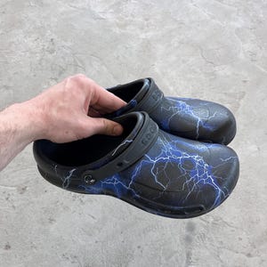 May include: Black clogs with a blue lightning bolt pattern. The shoes have a black strap and a molded design. The clogs are made of a rubber-like material and are shown on a gray surface.