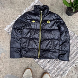 May include: A black puffer jacket featuring a high collar, a full-length front zipper with yellow trim, and two side pockets. The chest displays a yellow Kappa logo on the right and a yellow and black BVB 09 soccer club emblem on the left.