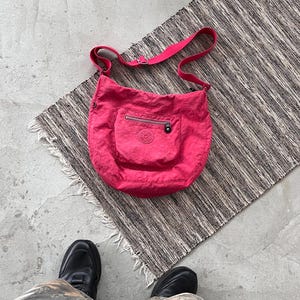 Op de afbeelding: Een felroze schoudertas met een voorvak en een ritssluiting. De tas heeft een bijpassende roze band en een cirkelvormig logo op het voorvak. De tas is gemaakt van een lichtgewicht materiaal.
