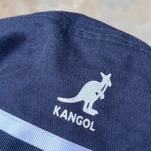May include: Navy blue Kangol hat with a white kangaroo logo and the brand name. The hat features two light blue stripes and is made of a textured fabric. The hat is a classic style.